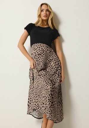 Femme portant un haut noir et une robe beige avec des pois noirs irréguliers, posant devant un mur uni de couleur claire.