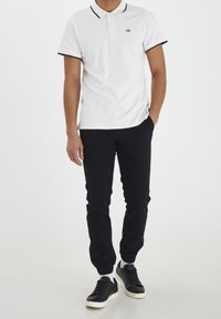Polo blanc avec des accents bleu marine, associé à un pantalon de jogging noir et des baskets noires. Texture lisse, coupe décontractée et coutures détaillées.
