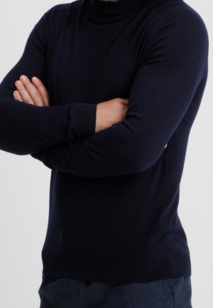 Mann trägt einen eng anliegenden schwarzen Rollkragenpullover und steht mit verschränkten Armen vor einem schlichten Hintergrund.