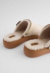 Pantoufles en shearling beige à enfiler avec une semelle en caoutchouc tan, dotées d'une large sangle et d'une semelle intérieure texturée. Design doux et confortable pour une utilisation intérieure.