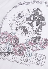 Camiseta blanca gráfica con un diseño de calavera en negro y gris, acompañado de rosas rosas y azules, con el texto "The Hoolies Established Since 2008".