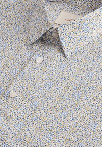 Lyseblå skjorte med knapper, der har et blomstermønster i gult og blåt, med en struktureret bomuldsfornemmelse og hvide knapper foran.