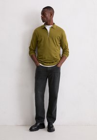 Olivgrünes Zip-up Langarmhemd über einem weißen T-Shirt, kombiniert mit schwarzen Jeans und schwarzen Schuhen, steht vor einer neutralen Wand.