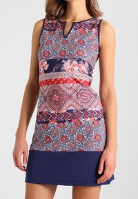 Kolorowa bezrękawnikowa sukienka z kwiatowymi i geometrycznymi wzorami w odcieniach czerwieni, granatu i bieli. Dekolt w kształcie biżuteryjnym oraz dopasowany krój z rozkloszowanym dołem.