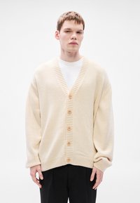 Bézs színű, V-nyakú kötött kardigán bordázott szegélyekkel és hat barna gombbal az elején, fehér póló fölött viselve.