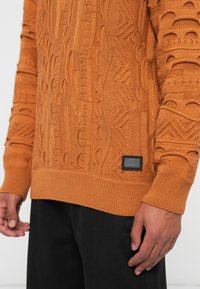Πλεχτό πορτοκαλί πουλόβερ με υφαντές σχέσεις, ριμπ μανσέτες και μαύρο λογότυπο στην κάτω αριστερή πλευρά. Φοριέται με μαύρο παντελόνι.