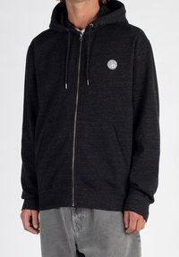 Black zip-up hoodie made of textured fabric with a front pocket, featuring a circular white logo on the left chest.