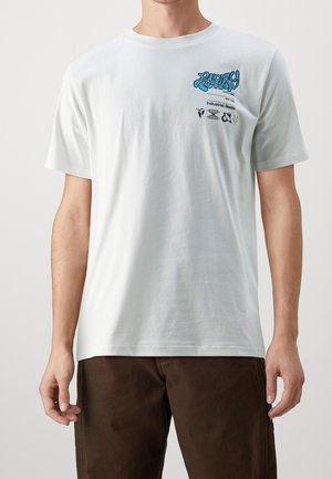 Man wearing white T-shirt with blue and black graphic text and icons on upper left chest, paired with dark brown pants.