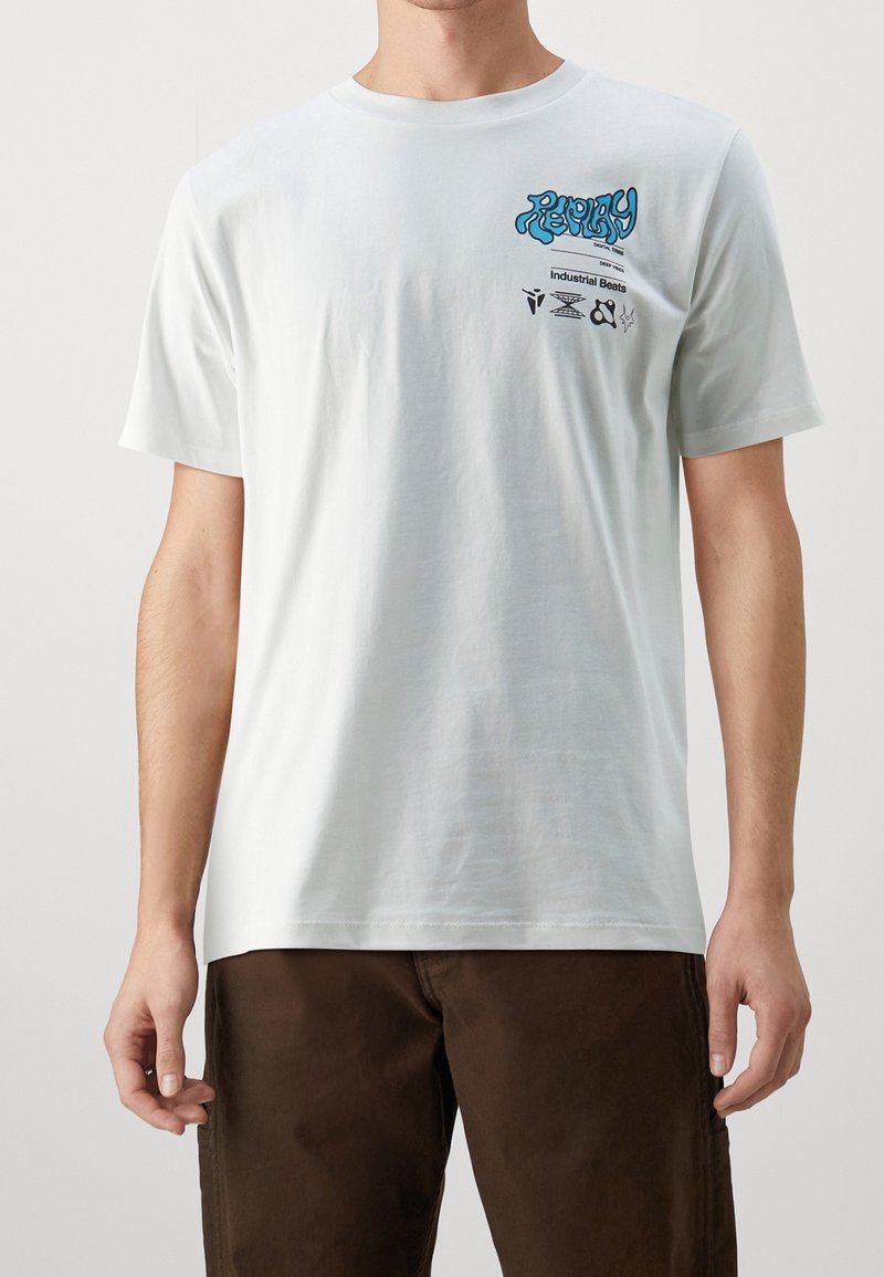 Man wearing white T-shirt with blue and black graphic text and icons on upper left chest, paired with dark brown pants.