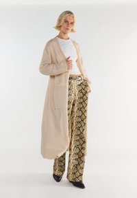 Blondynka w beżowym długim kardiganie, białej bluzce, szerokich spodniach w wężowy print i czarnych czółenkach, pozująca na białym tle.