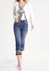 Veste blanche texturée, haut métallique argenté, jean bleu court avec accents de franges à l'ourlet, et ballerines argentées. Fond épuré.