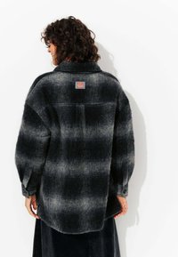Chemise en flanelle noire et grise à carreaux surdimensionnée avec une texture douce, des manches boutonnées, et un patch logo dans le dos.