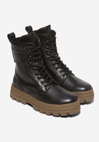 Paire de bottes de combat en cuir noir avec semelles en caoutchouc beige épaisses et lacets noirs sur fond blanc.