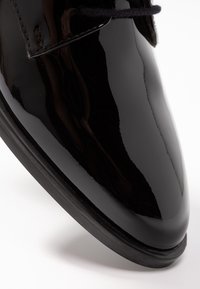 Chaussure de ville en cuir noir brillant avec lacets noirs et couture détaillée sur fond blanc.