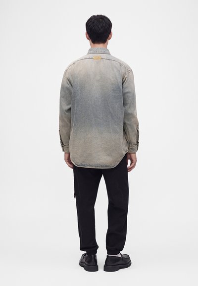 Homme debout de dos portant une chemise en jean grise délavée, un pantalon noir et des chaussures noires sur un fond clair uni.