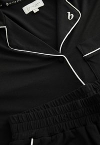 Fekete pizsama szett, rövid ujjú felsővel, fehér paszpól díszítéssel és mellzsebbel, valamint gumis derekú rövidnadrággal. Puha anyagtextúra.