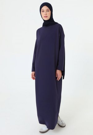 Donna che indossa un abito lungo a maniche lunghe blu navy con hijab abbinato e sneaker bianche, in piedi davanti a uno sfondo bianco semplice.