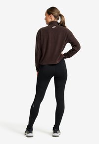 Frau mit brauner Fleecejacke und schwarzen Leggings, die mit einer Hand auf der Hüfte gegenüber einem einfarbigen Hintergrund steht.