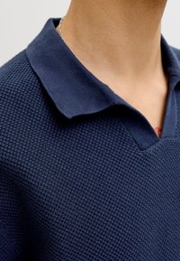 Gros plan d'une personne portant un polo bleu marine texturé avec un col replié et un collier chaîne en argent.