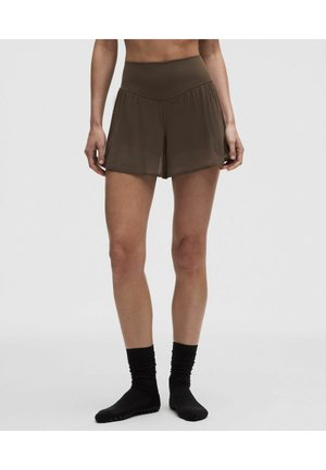 Persoon draagt hooggetailleerde bruine sportshort en zwarte gripkousen, staand op een effen lichtgrijze achtergrond.