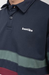 Marineblauwe polo met een tweeknoopssluiting, voorzien van horizontale strepen in groen en bordeaux. "Kaotiko" logo geborduurd op de borst.