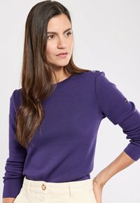 Lila Sweatshirt mit gerippter Textur, ausgestattet mit Knopfdetail an den Schultern und enganliegenden Ärmeln, kombiniert mit cremefarbigen Cordhosen.
