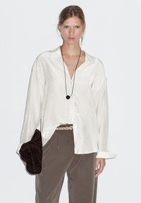 White satin shirt with a relaxed fit, long sleeves, and a collar, paired with beige pants and a dark brown clutch bag.