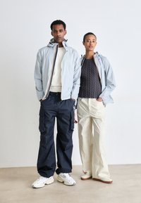Helehall zip-up jakk koos kapuutsiga, kontrastiks tumesinised cargo püksid ja kreemjad laiad püksid. Jalatsiteks on massiivsed spordijalatsid. Minimalistlik keskkond.