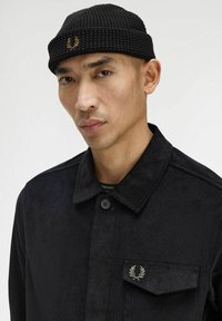 Giacca in corduroy nero con chiusura a bottoni, dotata di taschino sul petto con dettaglio ricamato a corona d'alloro e un berretto nero lavorato a maglia.