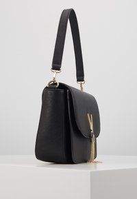 Bolso de hombro de cuero negro con solapa plegable, herrajes dorados y un detalle de borlas de cadena. Presenta una forma estructurada y una correa ajustable.