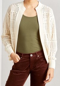 Pletený cardigan v slonovinové s otevřeným vzorem, žebrovaným límcem a manžetami, spárovaný s olivovým tílkem a hnědými kordovými kalhotami.