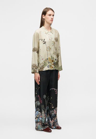 Blouse en soie vert pâle à imprimé floral ; associée à un pantalon large noir orné de motifs botaniques colorés et des chaussures bordeaux.