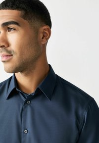 Mężczyzna z krótkimi ciemnymi włosami i przystrzyżoną brodą, ubrany w ciemnoniebieską koszulę zapinaną na guziki, patrzący w lewo na jednolity jasny tle.