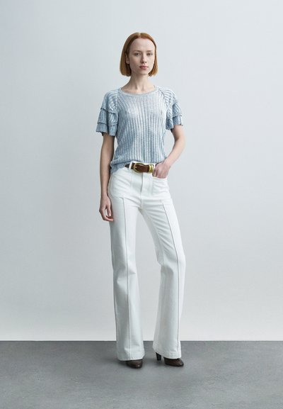 Haut en maille côtelée bleu clair avec manches à volants, associé à un pantalon blanc évasé. Une ceinture marron notable et des talons hauts complètent le look.