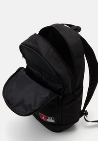Mochila negra hecha de tela duradera; cuenta con un gran compartimento principal con apertura con cremallera y bolsillo de malla; incluye un parche con el logo.