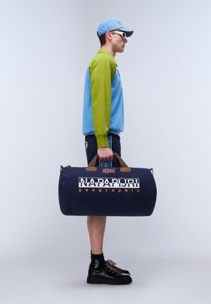 Uomo con giacca blu e verde, berretto blu e scarpe nere che porta una grande borsa da viaggio blu navy con il logo "Napapijri Geographic".