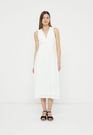 Kvinde stående iført en hvid midi-kjole uden ærmer med V-hals og sorte sandaler med ankelstrop mod en ensfarvet hvid baggrund.