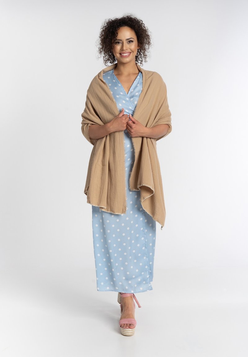 Jasnoniebieska sukienka w kropki z fasonem kopertowym, w połączeniu z beżowym, strukturalnym szalem. Modelka nosi różowe sandały na platformie.