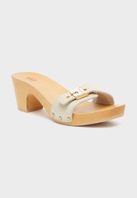 Beige leather slide sandal with a wooden wedge heel. Features a strap with gold hardware and decorative studs. Smooth texture.