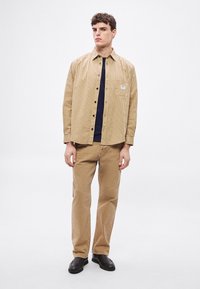 Srajca v bež barvi s črtami in gumbki ter z žepom na prsih, kombinirana s kaki hlačami in črnimi čevlji. Srajca ima sproščen kroj in mehko teksturo.