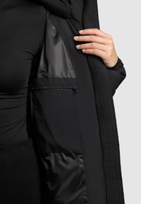 Interior de chaqueta de plumas negra, con forro suave, textura acolchada y un bolsillo con cremallera para almacenamiento.