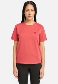T-shirt en coton rouge avec un col rond, des courtes manches, une coupe droite et un petit logo sombre sur la poitrine gauche. Porté avec des leggings noirs.
