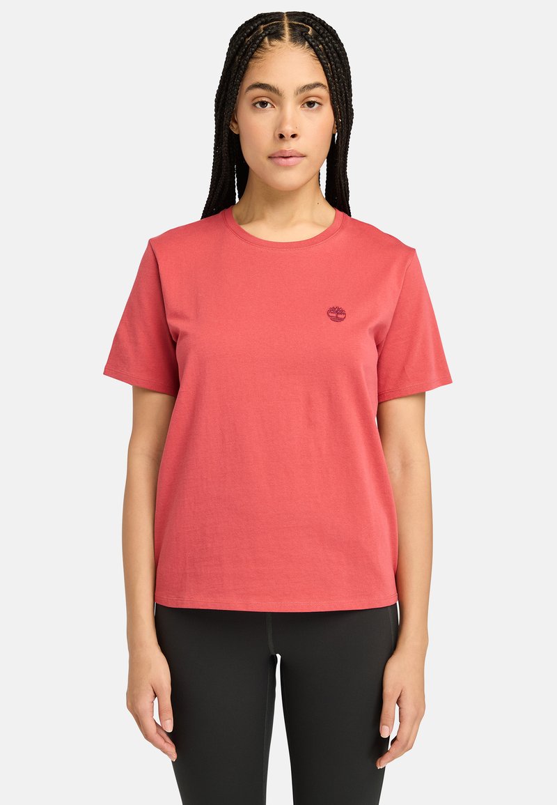 T-shirt en coton rouge avec un col rond, des courtes manches, une coupe droite et un petit logo sombre sur la poitrine gauche. Porté avec des leggings noirs.