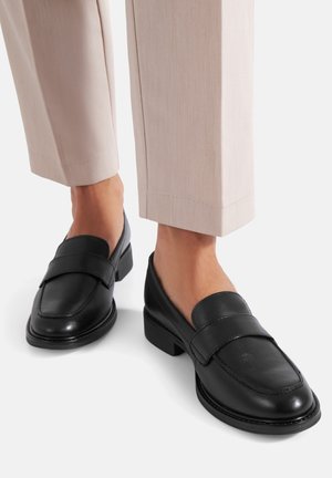 Zwarte lederen loafers met een lage blokhak, gedragen bij beige cropped broeken op een witte achtergrond.