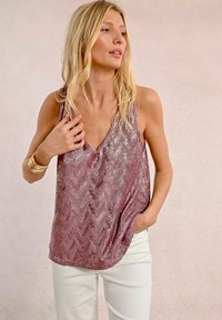 Top in paillettes viola con scollatura a V, vestibilità rilassata e motivo a chevron, abbinato a pantaloni bianchi. Texture liscia e lucentezza visibile.