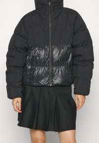 Svart puffjacka med hög krage, dragkedja och kontrasterande blänkande nederdel, med strukturerad yta och elastiska ärmslut.