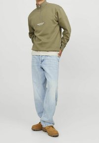 Olivgrön kvartslång zipper sweatshirt med vit logotyp, lager på en beige skjorta; parat med ljusblå lösa jeans och bruna skor.