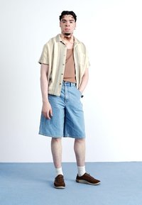 Kurzärmeliges gestreiftes Hemd in Beige und Hellgelb, braches Tanktop, blaue Jeansshorts, braune Turnschuhe und weiße Socken.