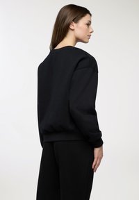 Donna con lunghi capelli castani che indossa una felpa nera oversize e pantaloni neri coordinati, rivolta di lato contro uno sfondo semplice.