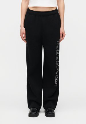 Γυναίκα που φοράει μαύρο παντελόνι με φαρδιά μπατζάκια και κάθετο λευκό κείμενο στο δεξί πόδι, συνδυασμένο με μαύρα παπούτσια και λευκό κοντό τοπ.